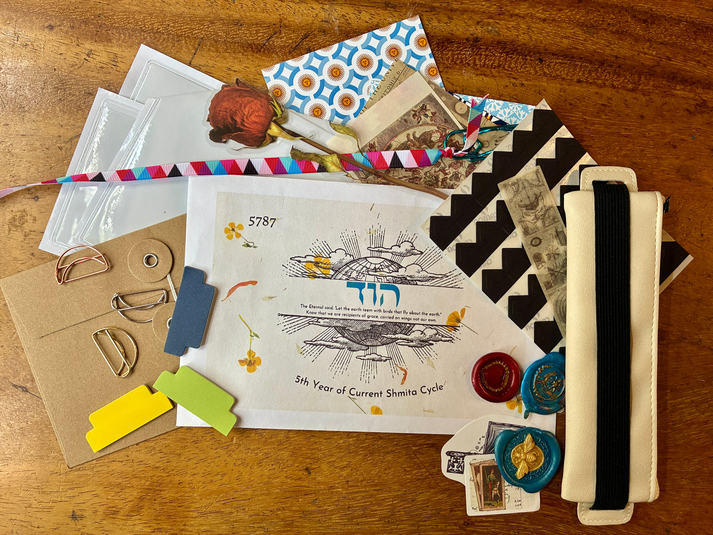 A flat-lay on a wooden table, featuring a central printed paper with Hebrew and English text, a black-and-white patterned envelope, a cream fabric pouch with a black elastic band, small colored paper pieces, metal rings, wax seals, and other ritual accessories.