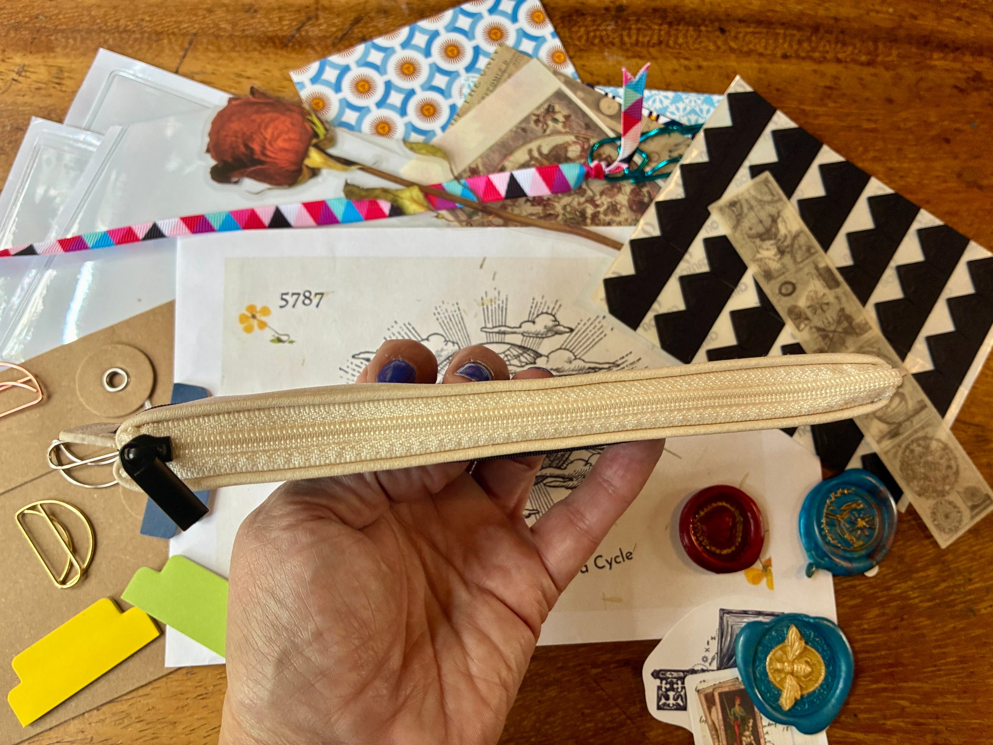A hand holding a beige zippered pouch with a textured pattern, surrounded by various colorful craft supplies, patterned papers, and wax seals on a wooden table.