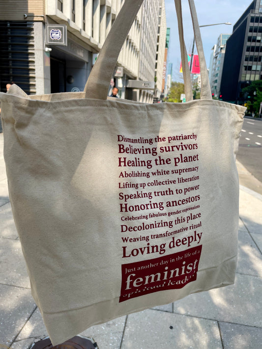 Off white tote bag with red text on a city street with feminist spiritual leader message